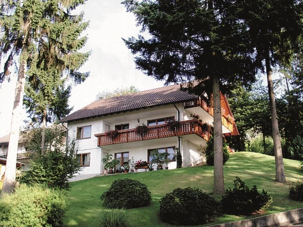 Ein zweistöckiges weißes Haus mit braunem Dach und großem Holzbalkon. Im Vordergrund eine Rasenfläche mit Nadelbäumen