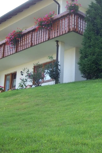 Ein zweistöckiges weißes Haus mit braunem Dach und großem Holzbalkon. Im Vordergrund eine Rasenfläche mit Nadelbäumen