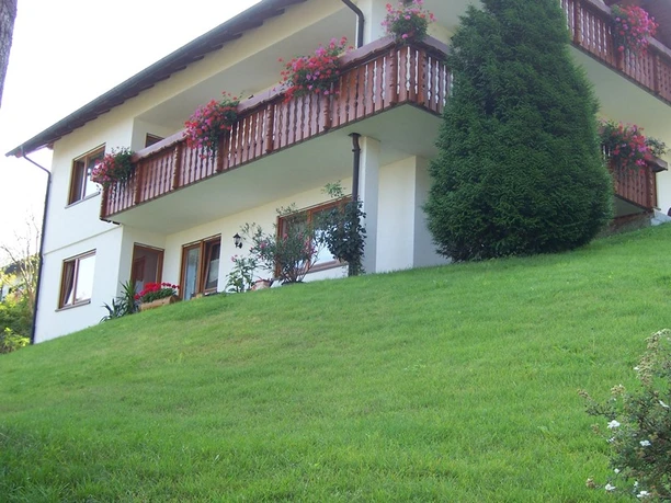 Ein zweistöckiges weißes Haus mit braunem Dach und großem Holzbalkon. Im Vordergrund eine Rasenfläche mit Nadelbäumen