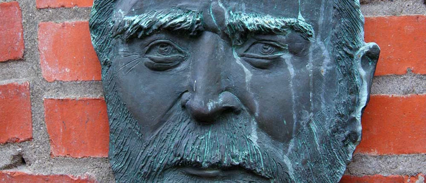 Gedenktafel Riedemann Bronzene Reliefbüste eines bärtigen Mannes an einer roten Backsteinwand.