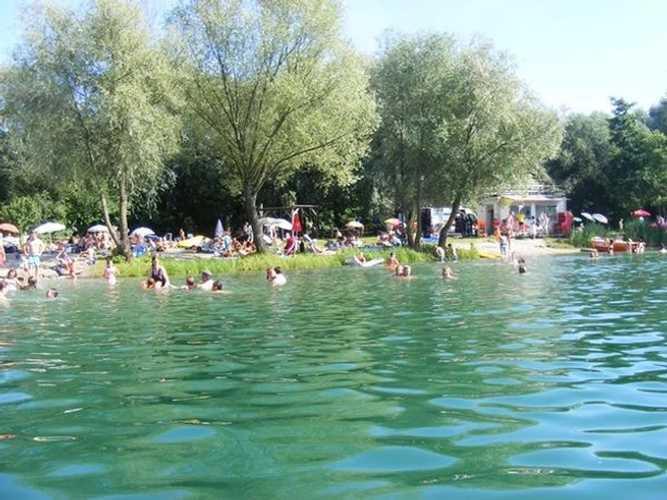 Badesee mit türkisfarbenem Wasser und Blick auf die Liegewiese mit Menschen in und am See