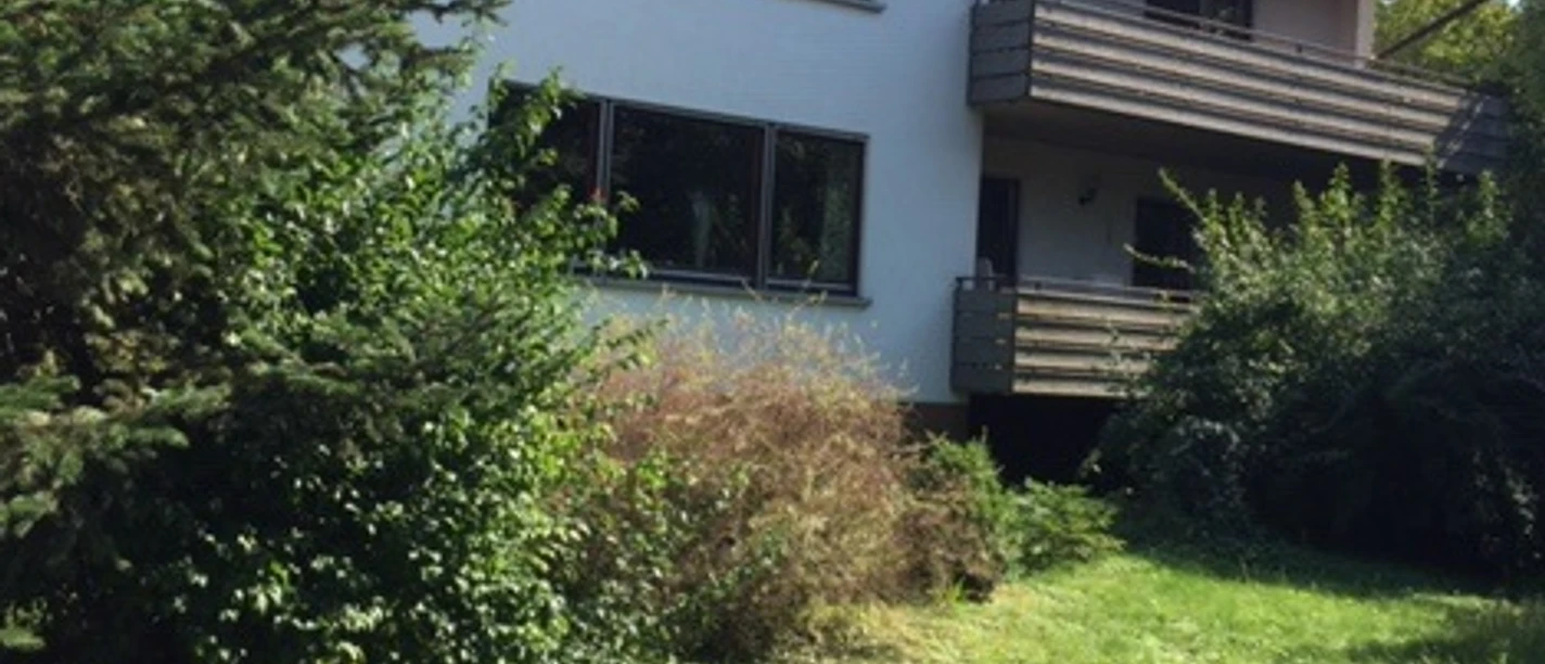 Außenansicht, weiße Hausfassade und zwei Balkone, davor Garten mit Büschen