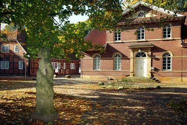 Kunstzentrum Koppelschleuse Backsteinbau mit Rundbogenfenstern und weißer Tür, umgeben von Bäumen und einer Steinskulptur im Hof.