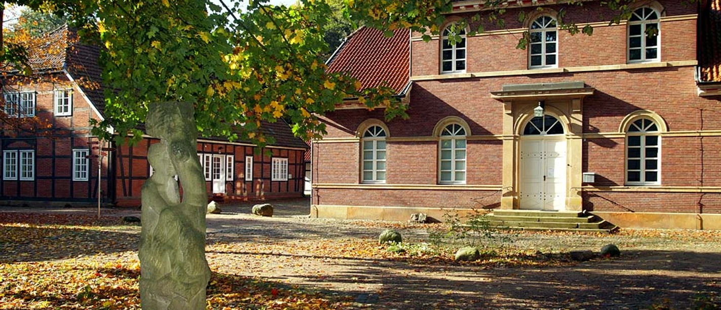 Kunstzentrum Koppelschleuse Backsteinbau mit Rundbogenfenstern und weißer Tür, umgeben von Bäumen und einer Steinskulptur im Hof.