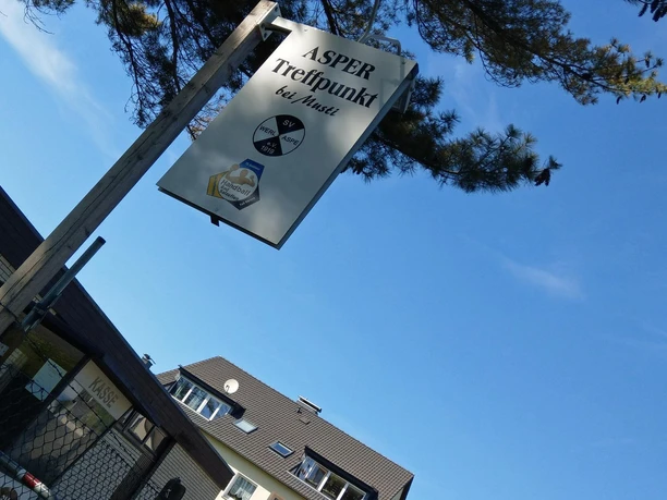 Schild des Asper Treffpunkts bei Musti vor blauem Himmel und Baumkronen