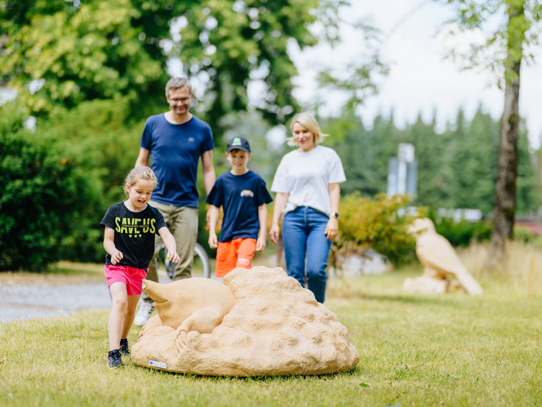Familie spaziert durch den Park, Kinder spielen fröhlich mit einem großen Kunstobjekt aus Stein.