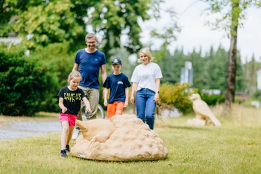 Tiere aus unseren Gärten und Siedlungen_besimmazhiqi-166.jpg Familie spaziert durch den Park, Kinder spielen fröhlich mit einem großen Kunstobjekt aus Stein.