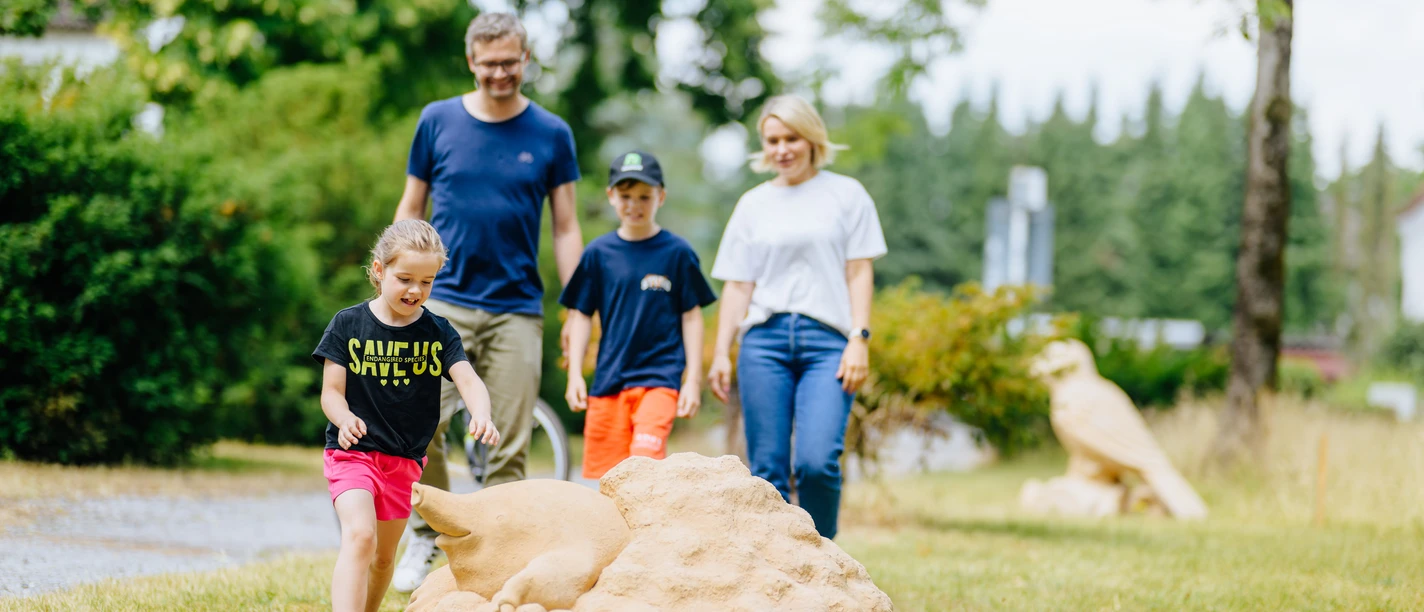 Tiere aus unseren Gärten und Siedlungen_besimmazhiqi-166.jpg Familie spaziert durch den Park, Kinder spielen fröhlich mit einem großen Kunstobjekt aus Stein.
