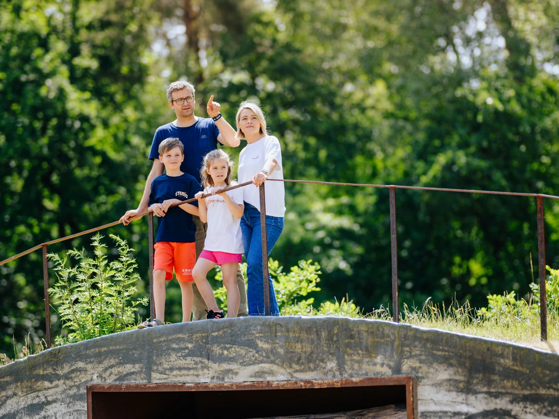 Familie auf einem bewachsenen, hölzernen Unterschlupf inmitten eines Waldes, blickt lächelnd umher.