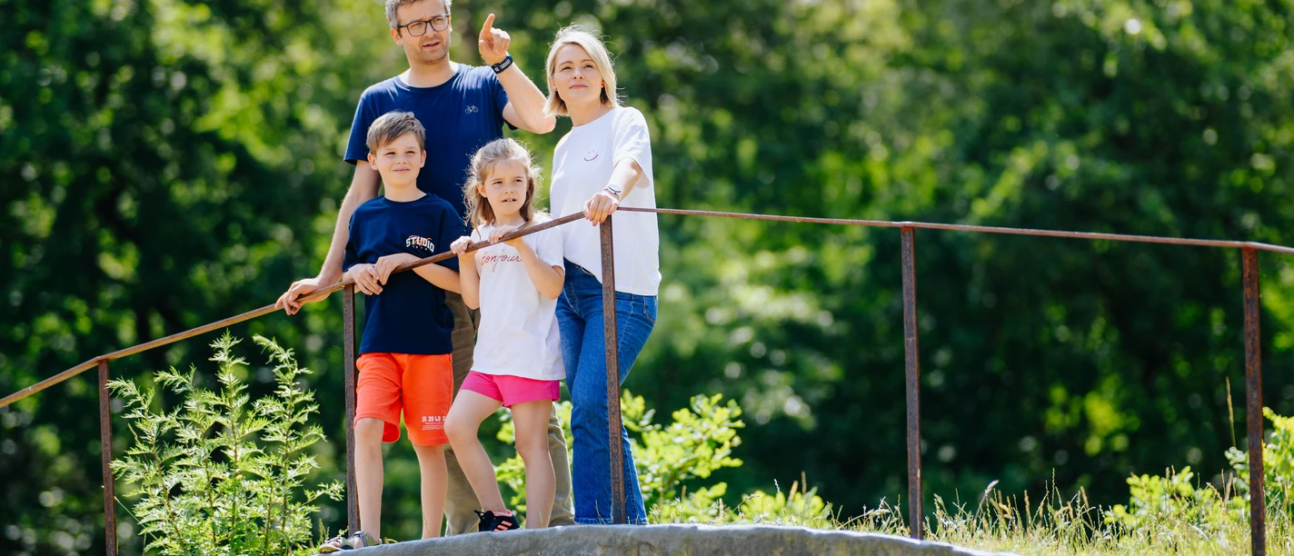 Familie auf einem bewachsenen, hölzernen Unterschlupf inmitten eines Waldes, blickt lächelnd umher.