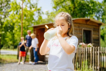 Der Alleenhof – ein Demeter-Bauernhof_besimmazhiqi-155.jpg Ein Mädchen trinkt Milch aus einer Flasche, während im Hintergrund eine Familie vor einer Holzhütte steht.
