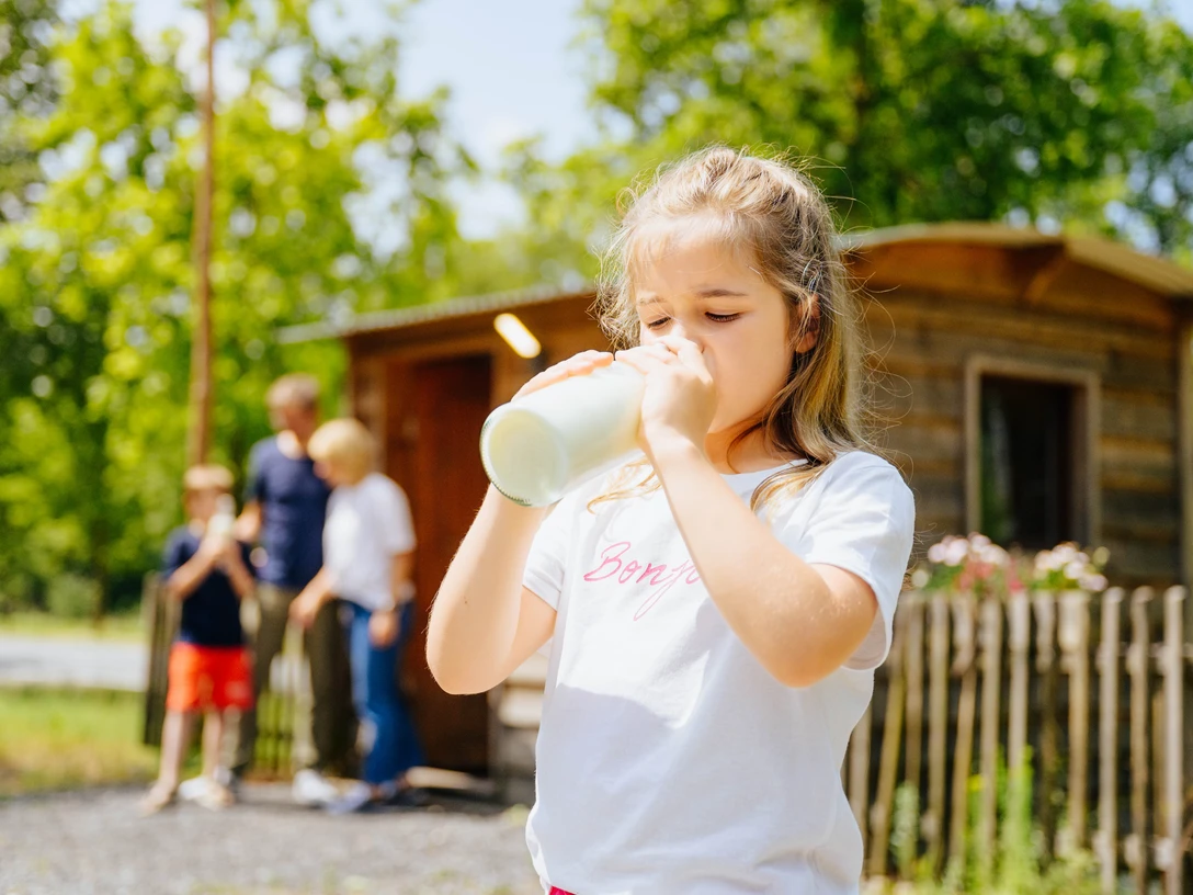 Der Alleenhof – ein Demeter-Bauernhof_besimmazhiqi-155.jpg Ein Mädchen trinkt Milch aus einer Flasche, während im Hintergrund eine Familie vor einer Holzhütte steht.