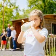 Der Alleenhof – ein Demeter-Bauernhof_besimmazhiqi-155.jpg Ein Mädchen trinkt Milch aus einer Flasche, während im Hintergrund eine Familie vor einer Holzhütte steht.