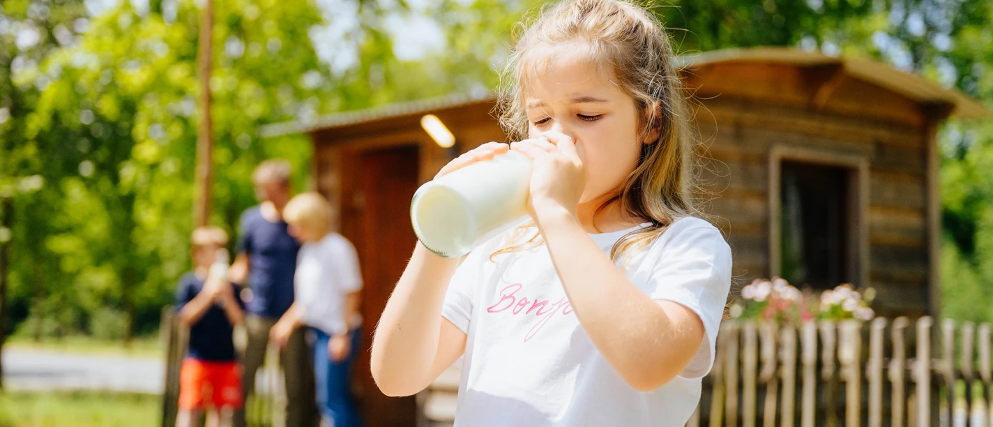 Der Alleenhof – ein Demeter-Bauernhof_besimmazhiqi-155.jpg Ein Mädchen trinkt Milch aus einer Flasche, während im Hintergrund eine Familie vor einer Holzhütte steht.