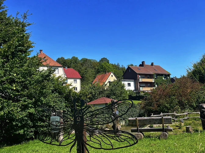 Eine Metallskulptur einer Libelle steht auf einer grünen Wiese, umgeben von Häusern und Bäumen.