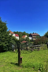 Fotospot Libelle_ Niklas Hecker Eine Metallskulptur einer Libelle steht auf einer grünen Wiese, umgeben von Häusern und Bäumen.