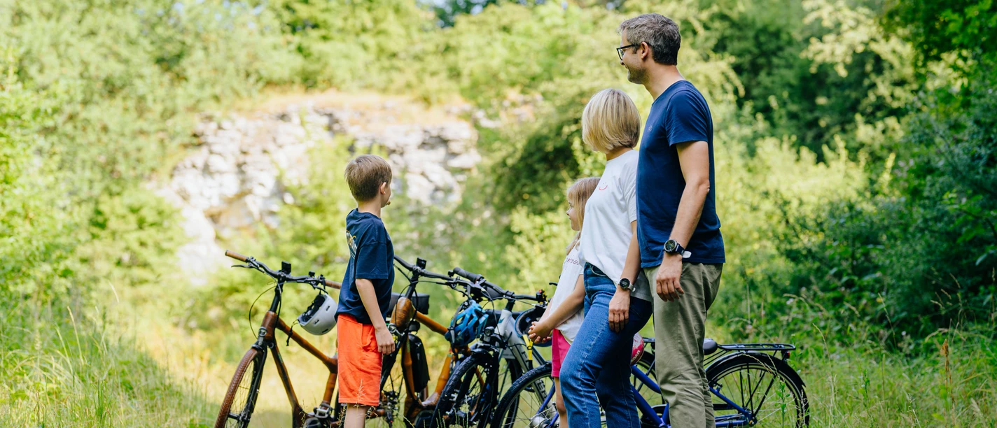 Familie mit zwei Kindern steht mit Fahrrädern in einem grünen Tal nahe eines alten Steinbruchs.