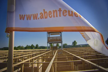 abenteuer-labyrinth-fahne De foto toont een witte vlag met op de voorgrond de inscriptie "www.abenteuer...". Op de achtergrond is een groot houten labyrint te zien, met daarop een hoge uitkijktoren.