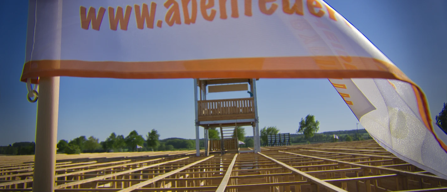 abenteuer-labyrinth-fahne The picture shows a white flag with the inscription "www.abenteuer..." in the foreground. A large wooden labyrinth can be seen in the background, topped by a high observation tower.