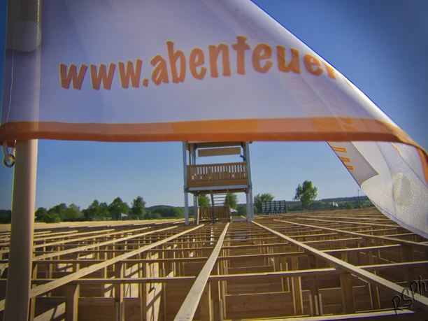 abenteuer-labyrinth-fahne Das Bild zeigt eine weiße Fahne mit der Aufschrift "www.abenteuer..." im Vordergrund. Im Hintergrund ist ein großes Labyrinth aus Holz zu sehen, über dem ein hoher Aussichtsturm steht.