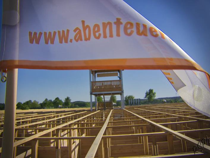 abenteuer-labyrinth-fahne Das Bild zeigt eine weiße Fahne mit der Aufschrift "www.abenteuer..." im Vordergrund. Im Hintergrund ist ein großes Labyrinth aus Holz zu sehen, über dem ein hoher Aussichtsturm steht.