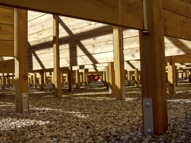 labyrinth-bispinger-heide The picture shows the view below a wooden hedge maze in the Bispinger Heide with pebbles on the ground, sunlight and shadows cast geometric patterns on the wooden construction.