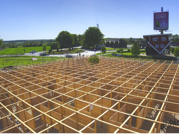 abenteuer-labyrinth-aussicht The picture shows a large wooden labyrinth from above. In the background are trees and a parking lot.