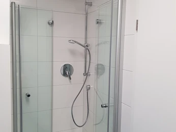 FeWo Liliencron 2 Moderne Duschkabine mit Glastüren, Regendusche und handlicher Brause vor weißen Fliesenwänden.