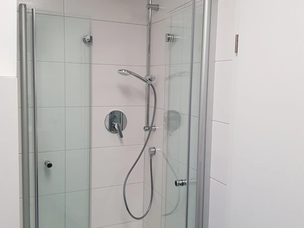FeWo Liliencron 2 Moderne Duschkabine mit Glastüren, Regendusche und handlicher Brause vor weißen Fliesenwänden.