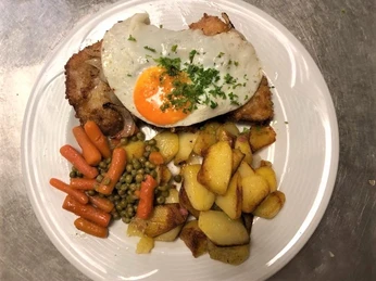 Schnitzel mit Spiegelei Ein gebratenes Schnitzel bedeckt mit Spiegelei, dazu Kartoffeln, Erbsen und Karotten auf weißem Teller.