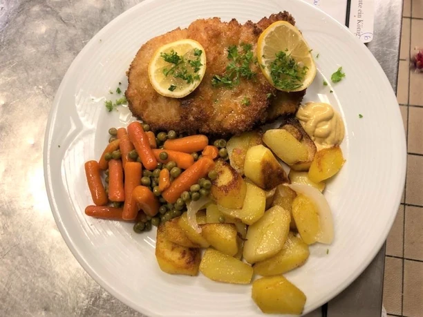 Schnitzel mit Zitronenscheiben, gebratenen Kartoffeln, Erbsen, Karotten und Senf auf einem weißen Teller.