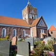 Ev.-ref. Kirche Pilsum Backsteinkirche in Pilsum mit hohem Vierungsturm unter blauem Himmel, umgeben von Friedhofsgräbern.