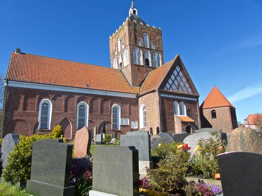 Ev.-ref. Kirche Pilsum Backsteinkirche in Pilsum mit hohem Vierungsturm unter blauem Himmel, umgeben von Friedhofsgräbern.