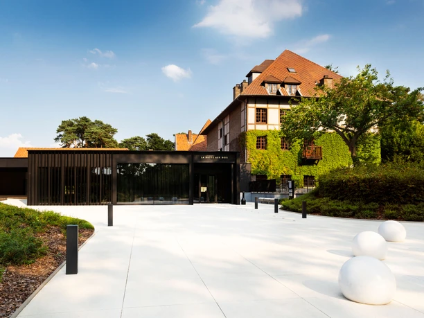 Vorplatz zu einem flachen, modernen Gebäude und einem großen Gebäude mit Fassadenbegrünung. Rechts und links sind bepflanzte Beete angelegt.