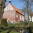 Kirche Upleward Backsteinkirche mit rotem Dach auf Warft, umgeben von Bäumen, Friedhof und schmalem Wassergraben.