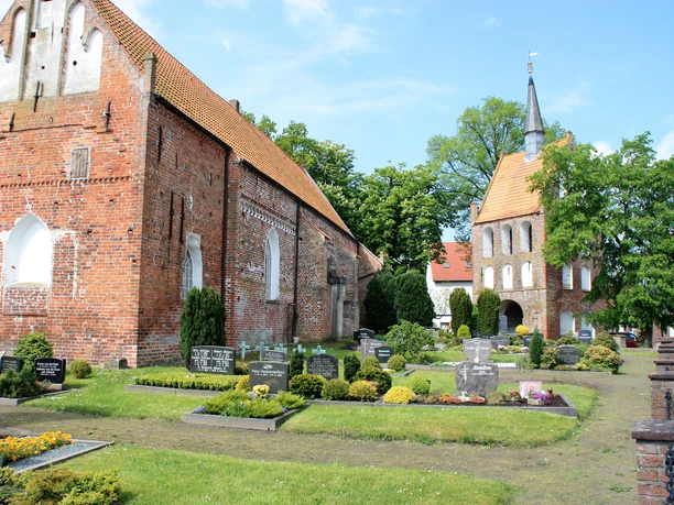 St. Nikolai Apen Historische Backstein-Kirche St. Nikolai in Apen mit gepflegtem Friedhof, umgeben von Bäumen am hellen Tag."