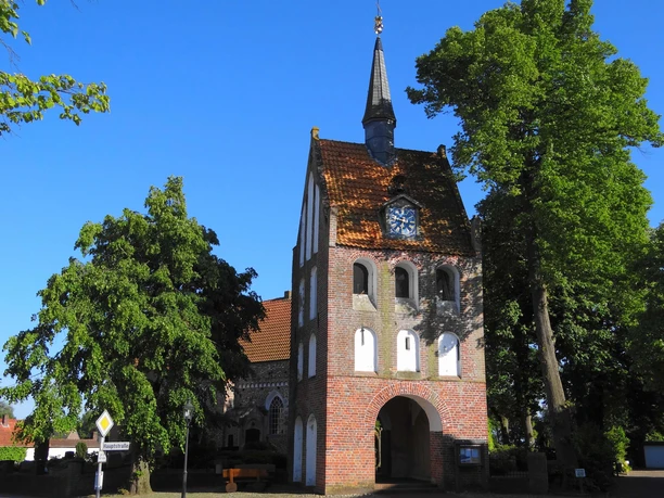 Ansicht des charmanten Backstein-Glockenturms von St. Nikolai in Apen, eingerahmt von grünen Bäumen.