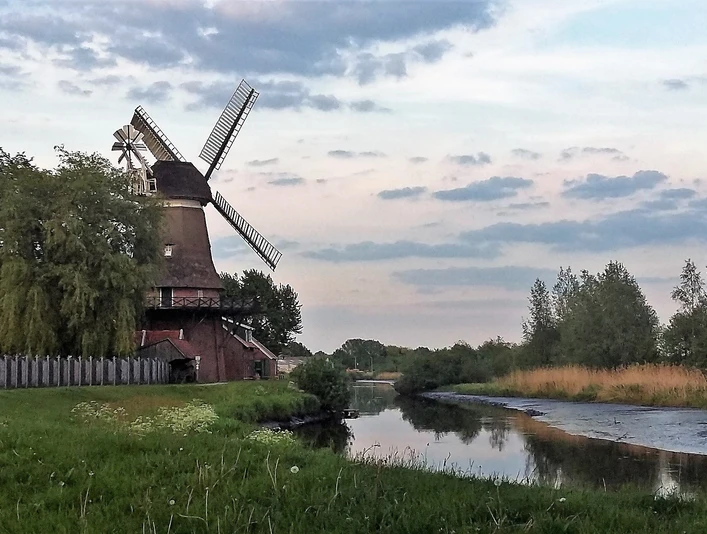 Hengstforder Mühle_Apen Die Hengstforder Mühle in Apen steht malerisch am Wasser, umgeben von üppigem Grünen unter blauem Himmel.