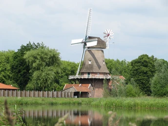 Hengstforder Mühle Die historische Hengstforder Mühle liegt malerisch an einem ruhigen Gewässer, umgeben von grünen Bäumen und roten Gebäuden. Eine klassische Windmühle mit weißen Flügeln und einem markanten, achteckigen Turm thront mittig im Bild, harmonisch in die ländliche Landschaft eingefügt.