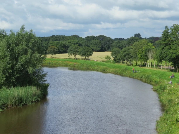 Ausblick von der Mühle Ein ruhiger Fluss schlängelt sich durch eine grüne Landschaft mit Wiesen und Bäumen unter einem bewölkten Himmel.