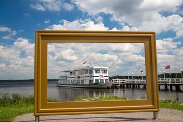 bilderrahmen-kurpark Ein großer Bilderrahmen steht im Vordergrund, durch den ein Fahrgastschiff auf dem Zwischenahner Meer sichtbar ist, mit Sonne und Wolken im Hintergrund.