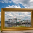 bilderrahmen-kurpark Ein großer Bilderrahmen steht im Vordergrund, durch den ein Fahrgastschiff auf dem Zwischenahner Meer sichtbar ist, mit Sonne und Wolken im Hintergrund.