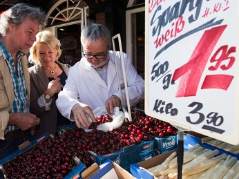 wochenmarkt-bad-zwischenahn Ein älterer Herr im weißen Kittel berät ein Paar beim Kauf von Kirschen auf einem Wochenmarkt.