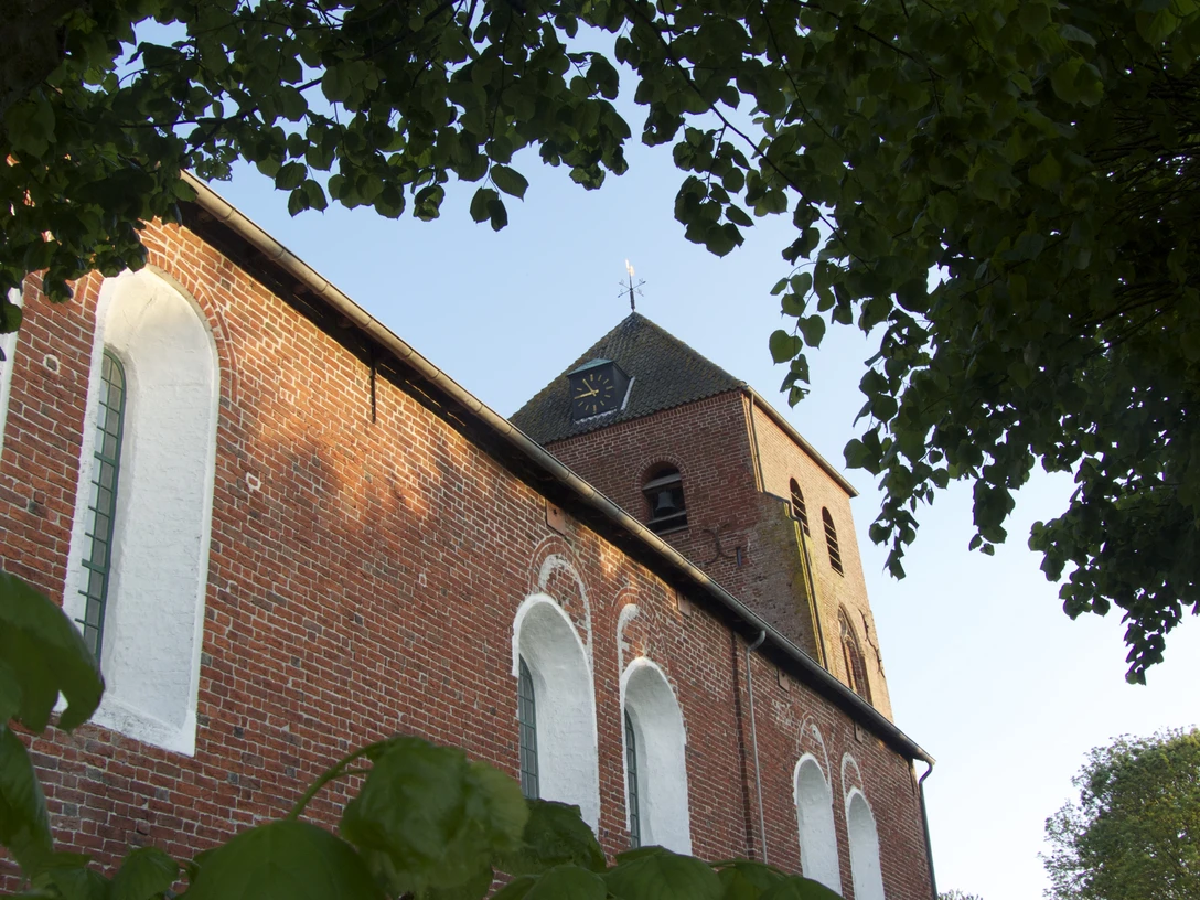 Ev.-ref. Kirche in Uttum Rote Backsteinkirche mit weiß abgesetzten Rundbogenfenstern und quadratischem Turm unter Bäumen.