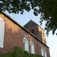 Ev.-ref. Kirche in Uttum Rote Backsteinkirche mit weiß abgesetzten Rundbogenfenstern und quadratischem Turm unter Bäumen.