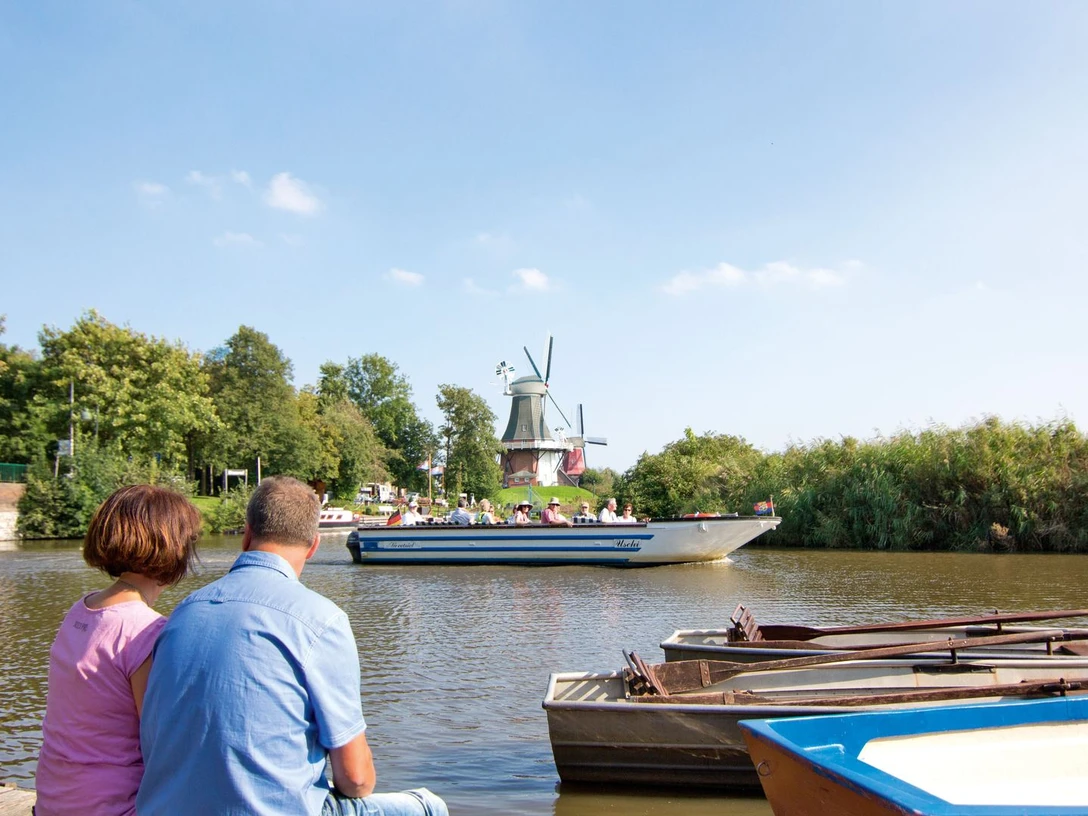 Abenteuer am Wasser Ein Paar sitzt am Wasser, im Hintergrund fährt ein Boot vor der Greetsieler Windmühle und grüner Landschaft.