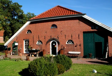 Atelier Glockenhaus_Erholungsort Rhauderfehn Rotes Backsteingebäude, Gockenhaus, mit rotem Ziegeldach, grüner Tür und mehreren kleinen Fenstern, davor Garten mit Sträuchern und Rasen