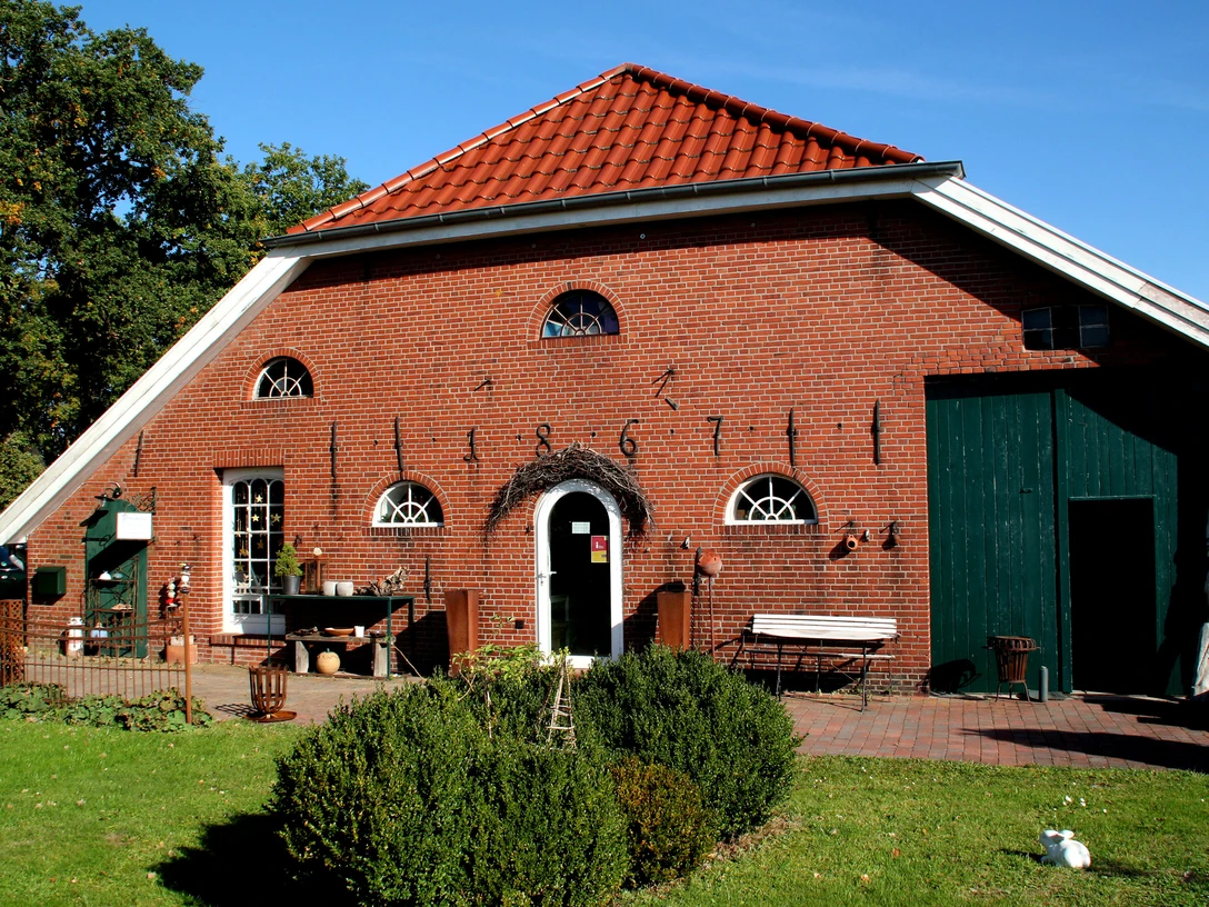 Atelier Glockenhaus_Erholungsort Rhauderfehn Rotes Backsteingebäude, Gockenhaus, mit rotem Ziegeldach, grüner Tür und mehreren kleinen Fenstern, davor Garten mit Sträuchern und Rasen