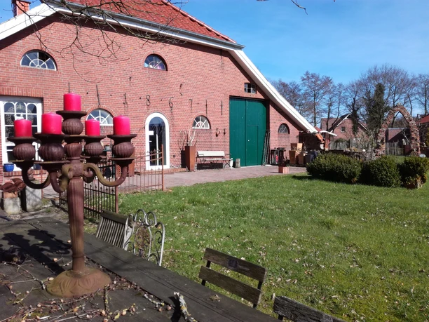 Atelier Glockenhaus_Erholungsort Rhauderfehn Rotes Backsteingebäude Atelier Glockenhaus mit grüner Tür und weißen Fenstern, davor ein großer Kerzenhalter mit roten Kerzen und Gartenmöbeln auf einer Wiese