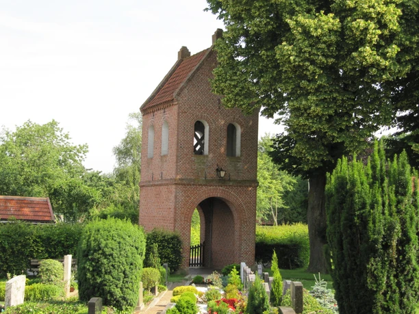 Backsteinglockenturm mit Bogendurchgang und Fenstern, umgeben von Bäumen und gepflegten Gräbern.